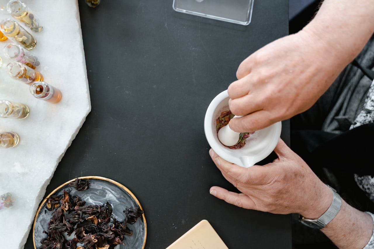 Hands preparing an herbal mixture using a mortar and pestle with dried flowers and oils.